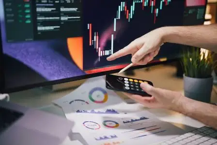 A cryptocurrency trader pointing at a candlestick chart on a monitor while holding a smartphone calculator over detailed financial P&L reports. Symbolizes the inadequacy of basic calculation tools for analyzing complex market returns and hidden fees.