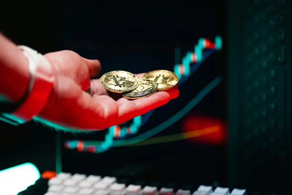 A person holding physical gold Bitcoin and Ethereum coins in their hand, with glowing cryptocurrency candlestick charts displayed in the background.